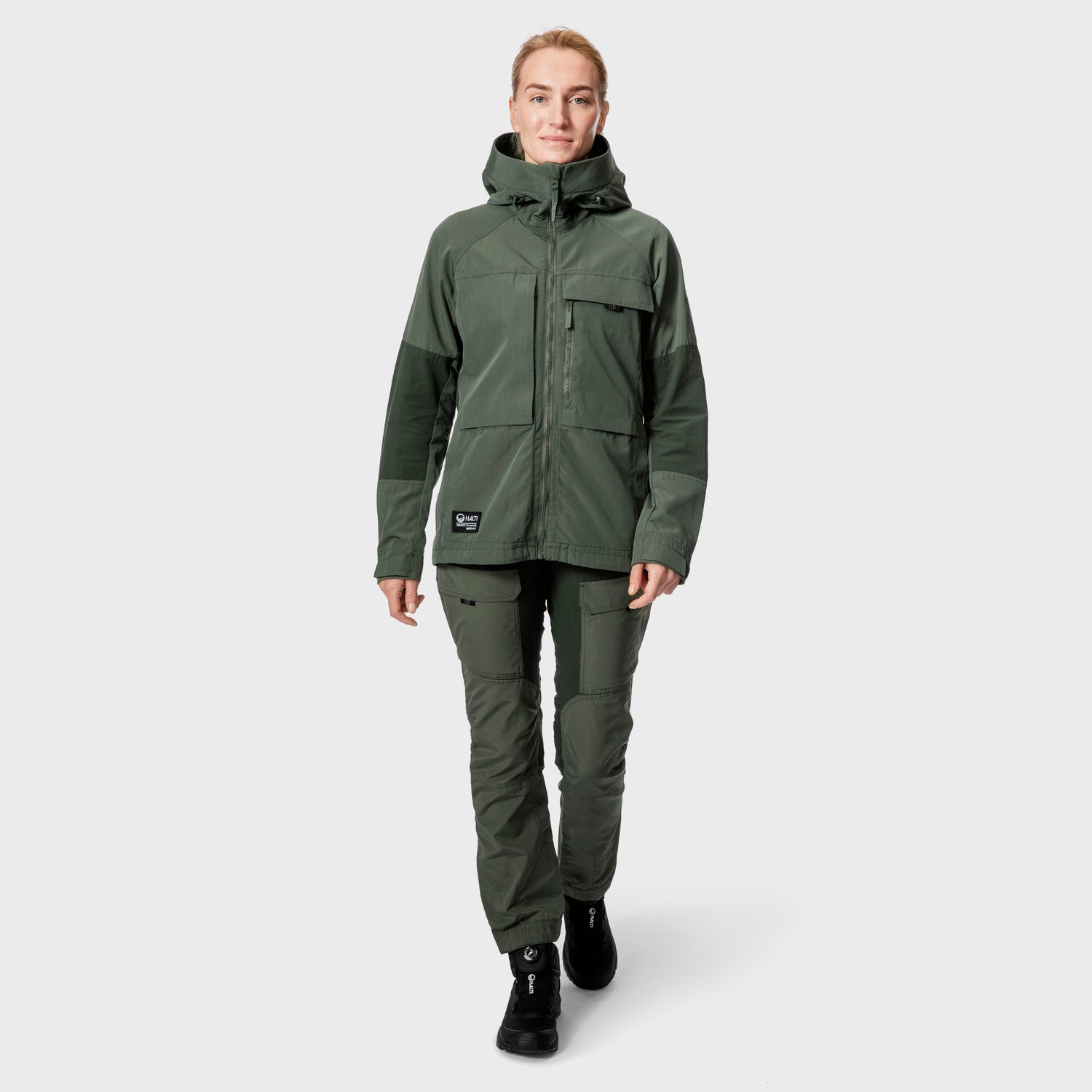 Halti Hiker Naisten Hengittävässä Retkeilytakissa ja hengittävästä kankaasta valmistetuissa housuissa oleva nainen seisoo harmaan taustan äärellä.