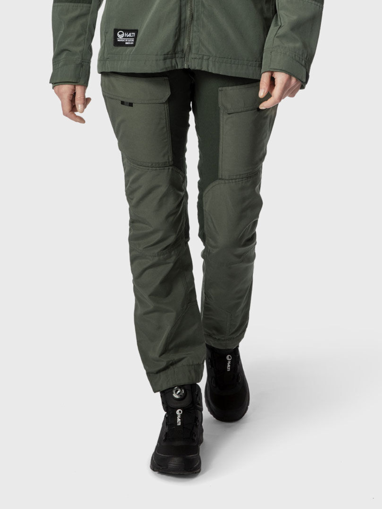 Henkilö Halti Hiker Naisten Hengittävissä retkeilyhousuissa, vihreissä ja mustissa vaelluskengissä, vyötäröstä alaspäin vaalealla taustalla.