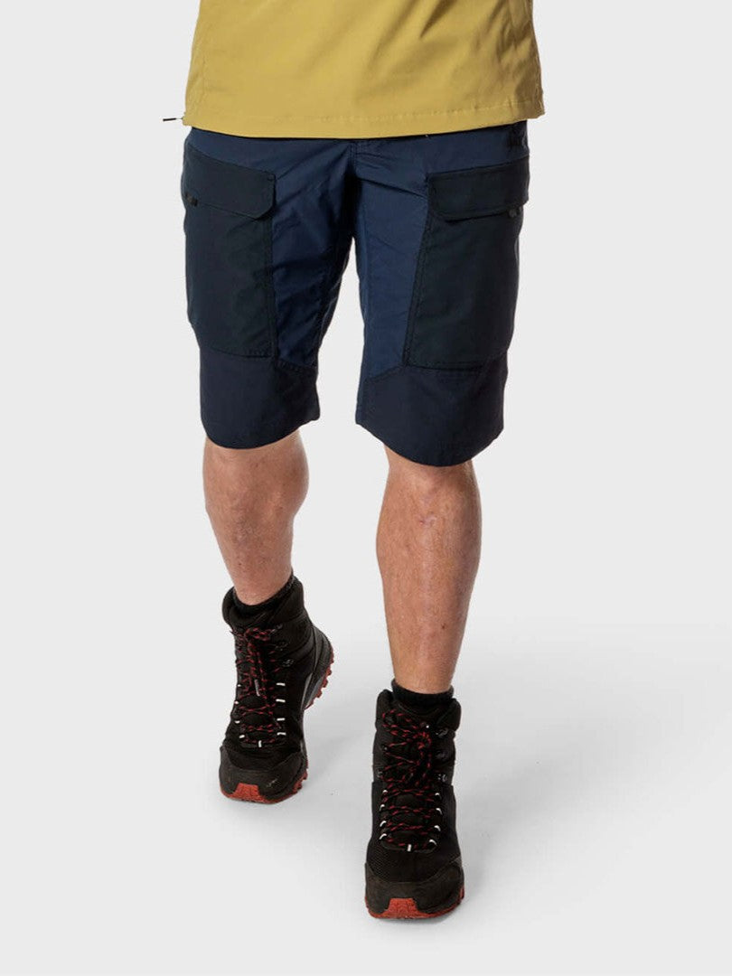 Henkilö, jolla on Halti Hiker Miesten Lite Shortsit, keltainen paita ja mustat vaelluskengät, seisoo tavallista taustaa vasten.