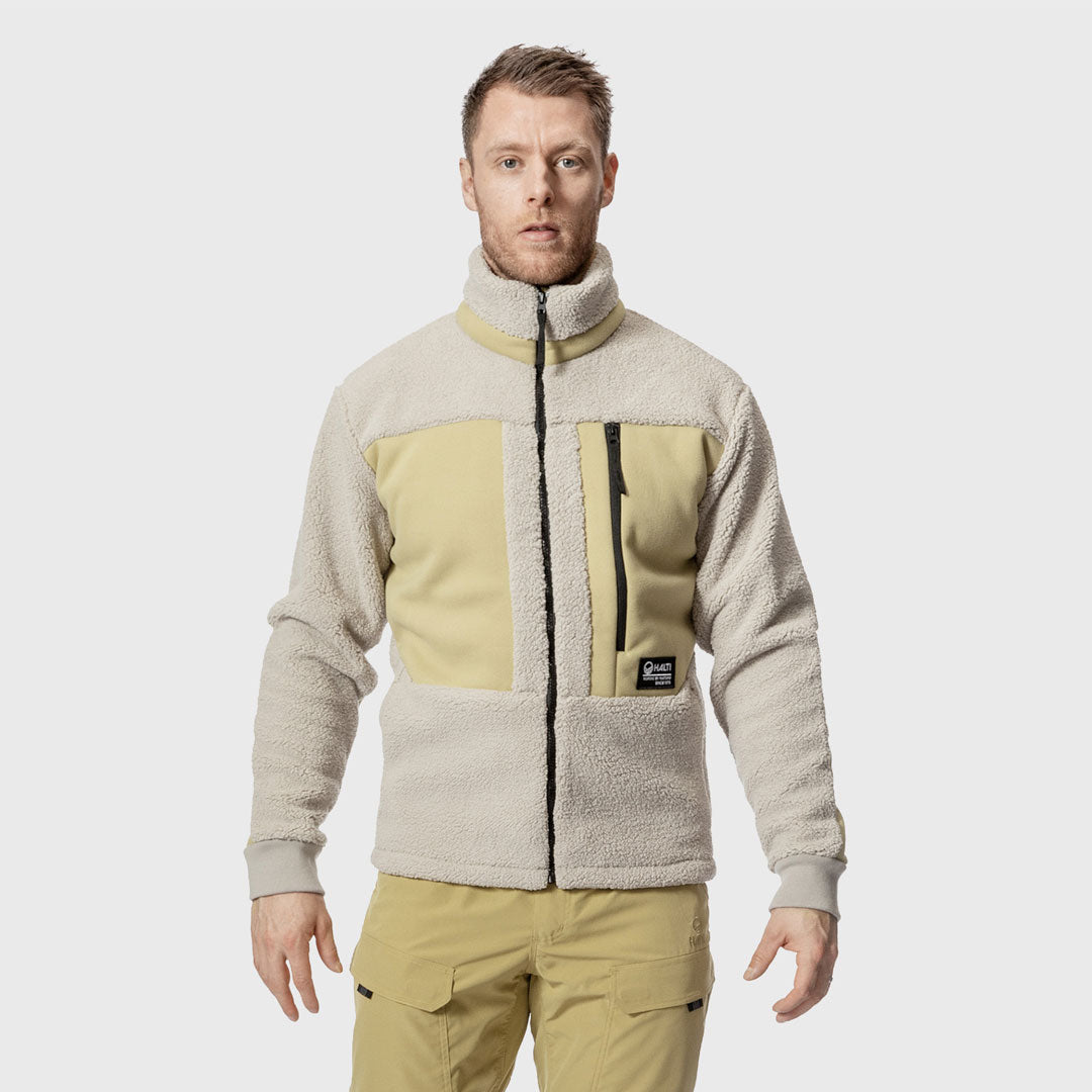 Mies seisoo kasvot menosuuntaan, yllään beige fleecetakki ja khakihousut valkoista taustaa vasten.