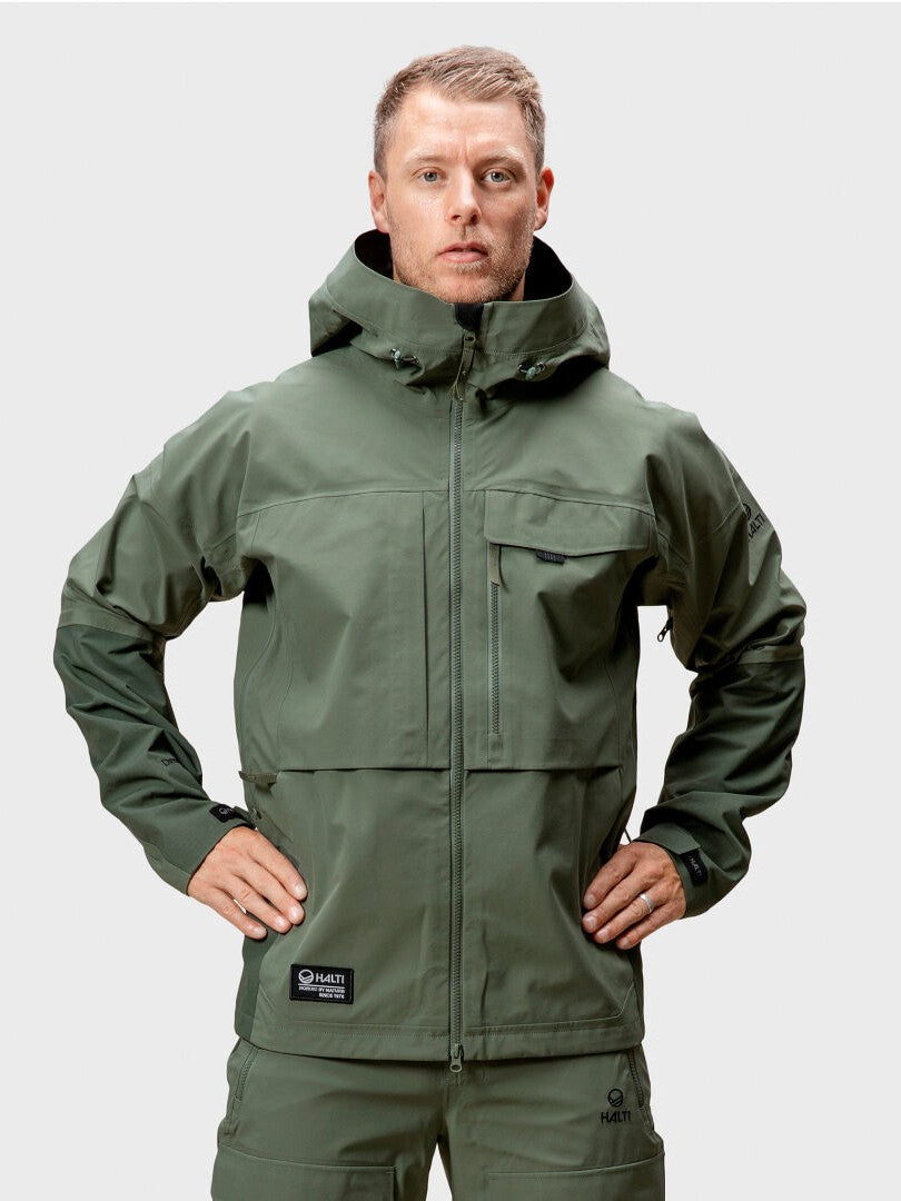 Mies katsoo eteenpäin vihreässä Halti Hiker DrymaxX 3L -tuulettuvassa takissa ja siihen sopivissa housuissa, kädet lantiolla, tavallinen tausta.