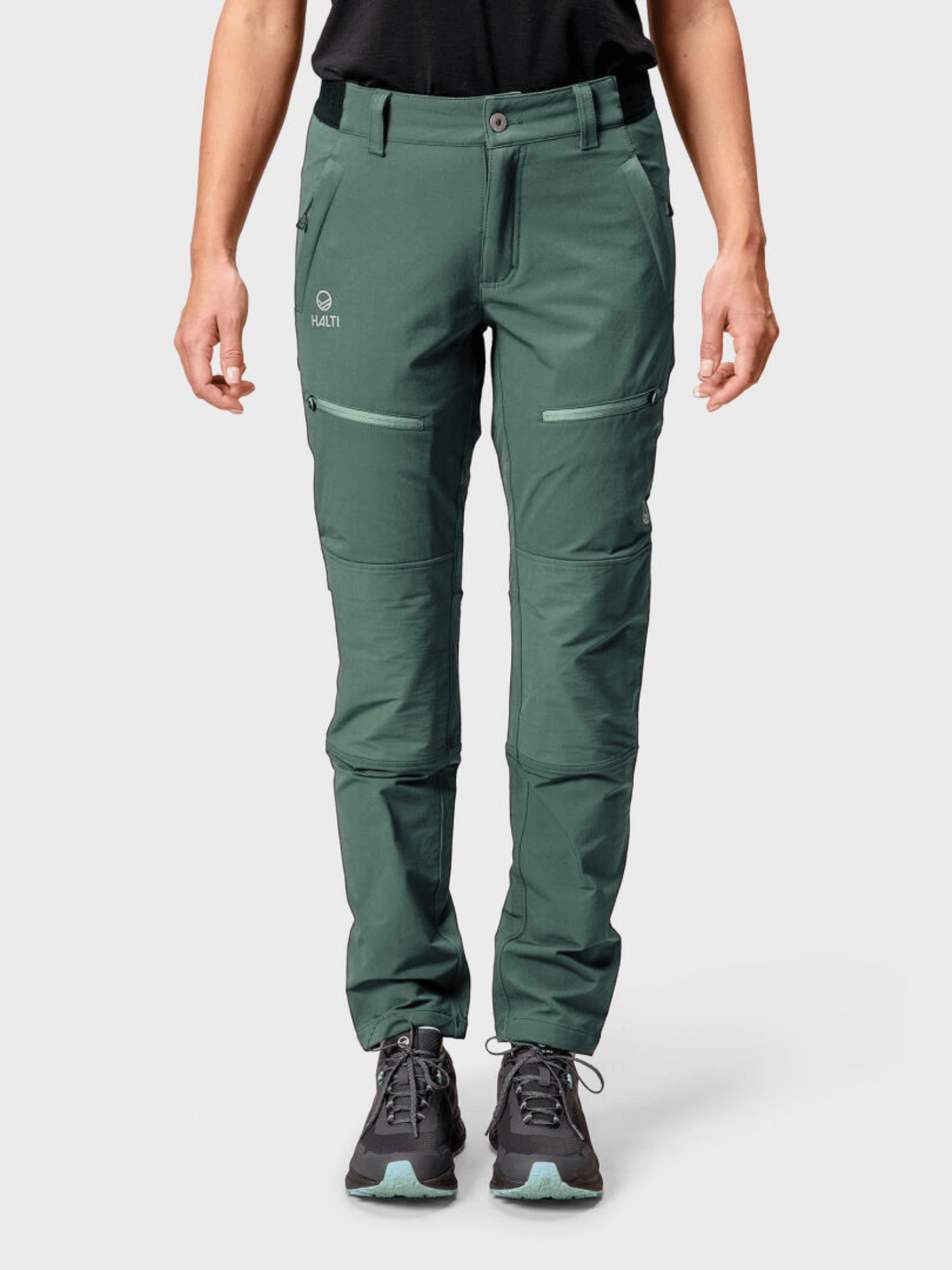 Henkilöllä on yllään vihreä Halti Pallas III Warm X-stretch Ulkoiluhousut Naiset ja mustat kengät, kasvot menosuuntaan harmaalla pohjalla.