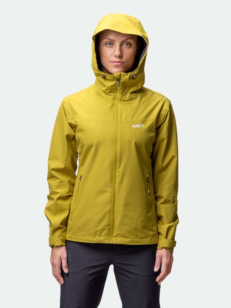 Henkilö, jolla on yllään keltainen Halti Forter DX Kuoritakki Naisten ja mustat housut, seisoo vaaleaa taustaa vasten.