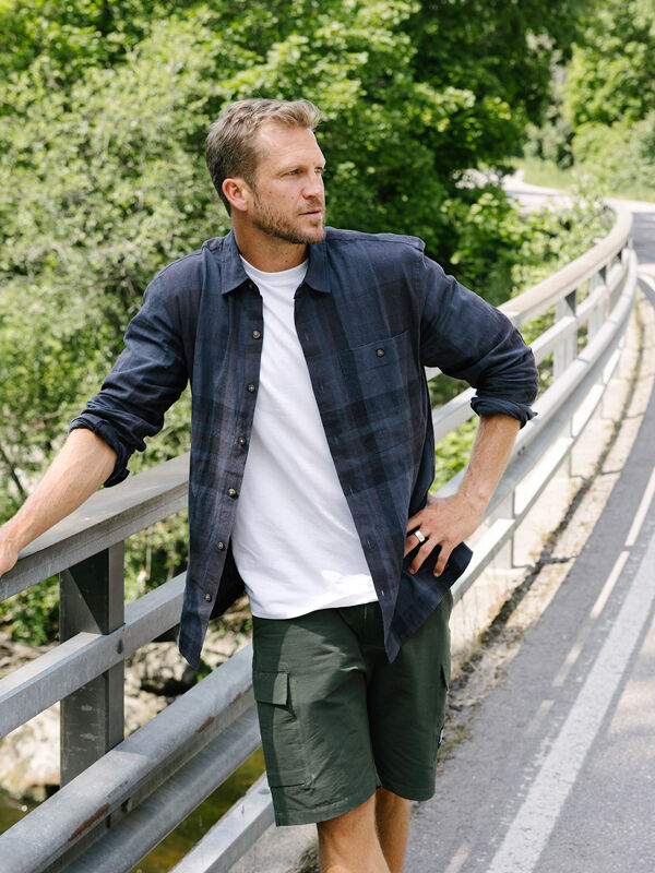 Mies Halti Hiker Retkeilypaita Miesten -takissa ja vihreissä shortseissa seisoo sillalla vehreyden takanaan.