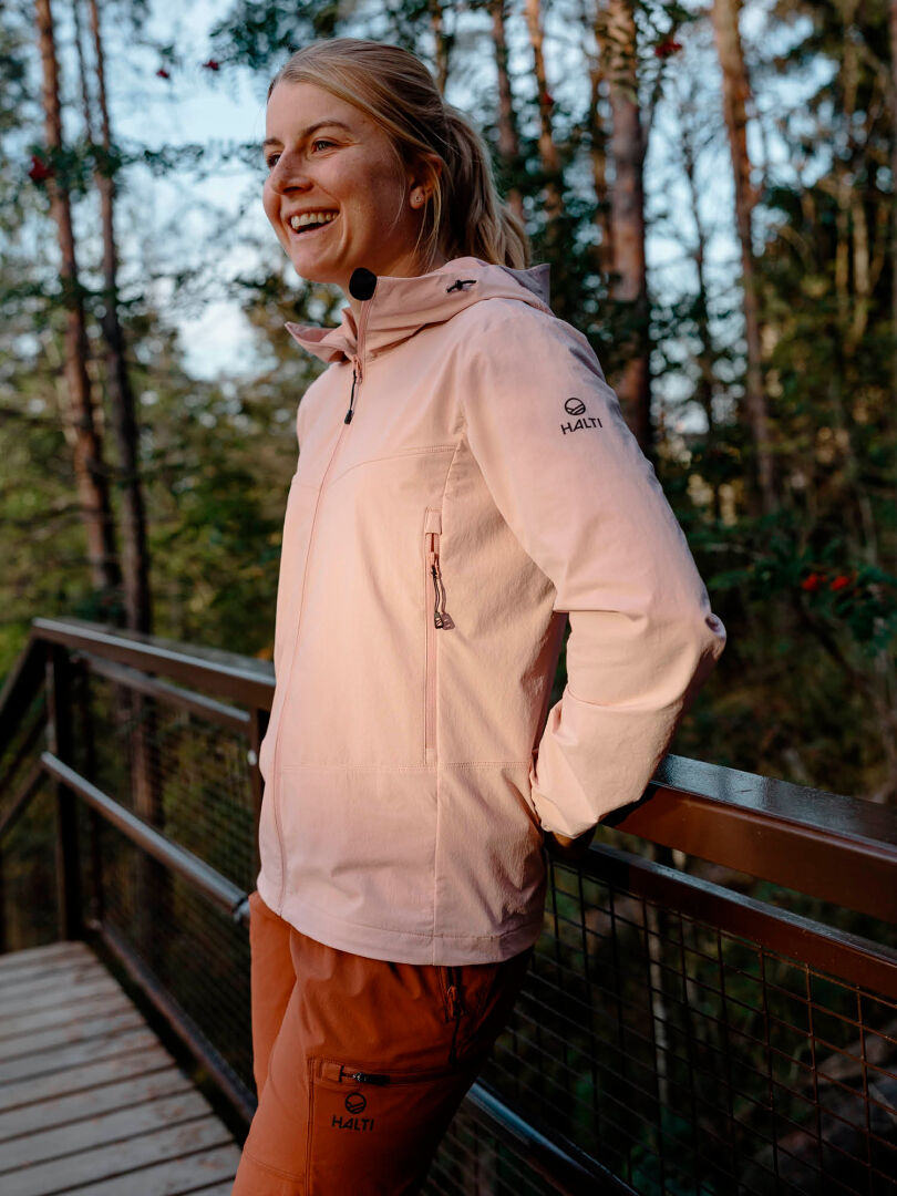 Vaaleanpunaisessa takissa ja Halti Pallas Evo X-stretch Housut Naisten -takissa oleva henkilö seisoo hymyillen puiden keskellä olevalla puisella sillalla.