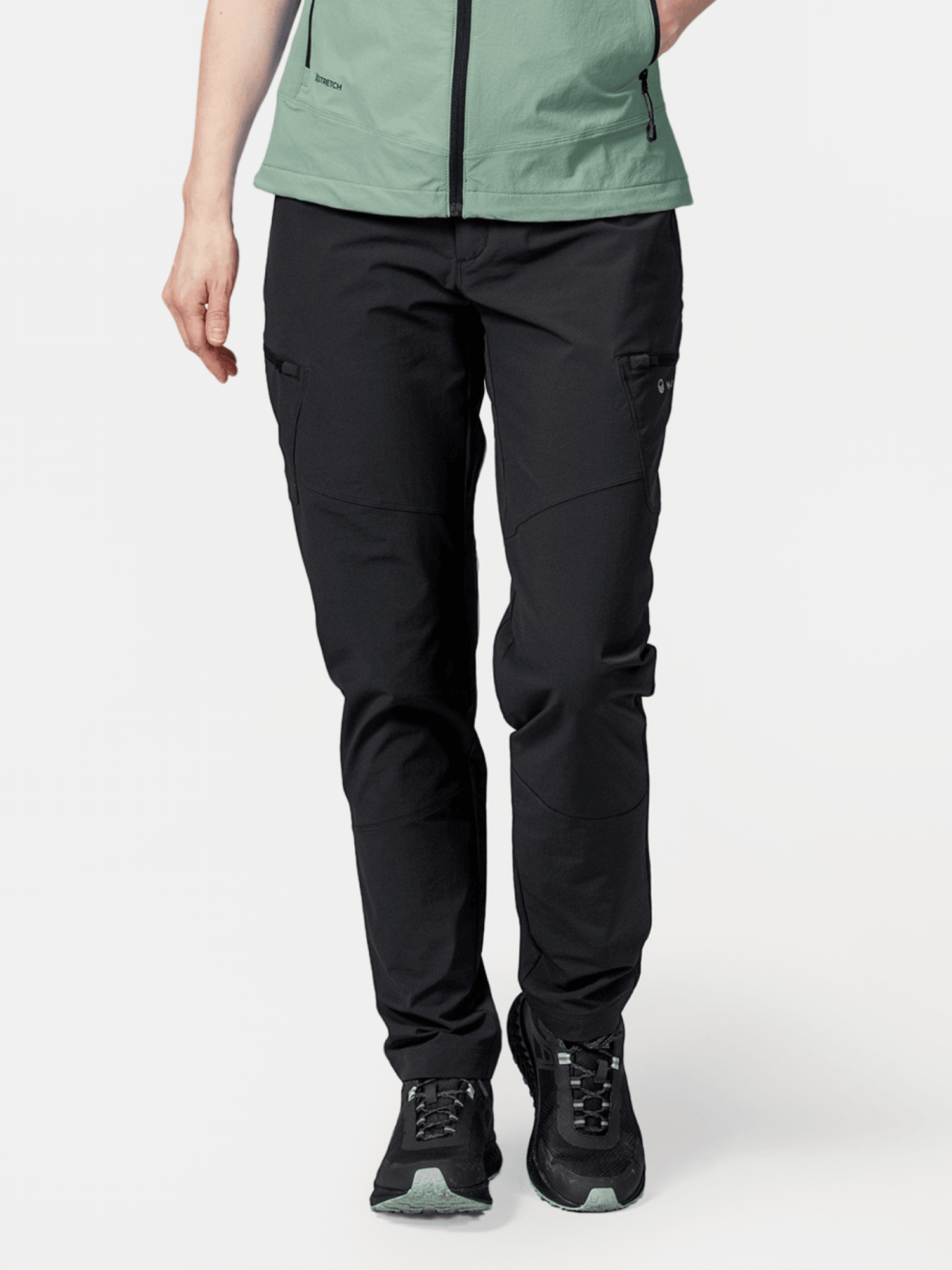 Henkilö, jolla on yllään mustat Halti Pallas Evo X-stretch -housut Naisille, vaaleanvihreä takki ja mustat lenkkarit valkoista taustaa vasten.