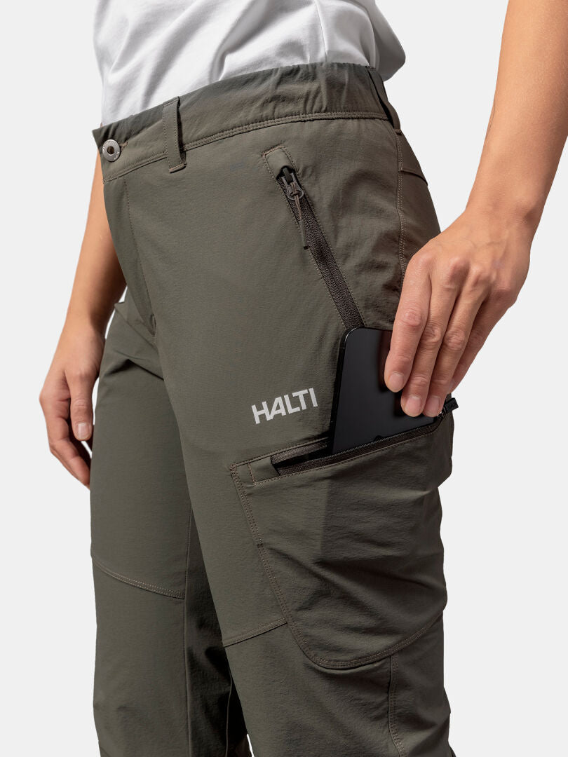 Henkilö laittaa mustan puhelimen oliivinvihreän Halti Pallas Evo X-stretch Housut Naisten vetoketjulliseen taskuun.