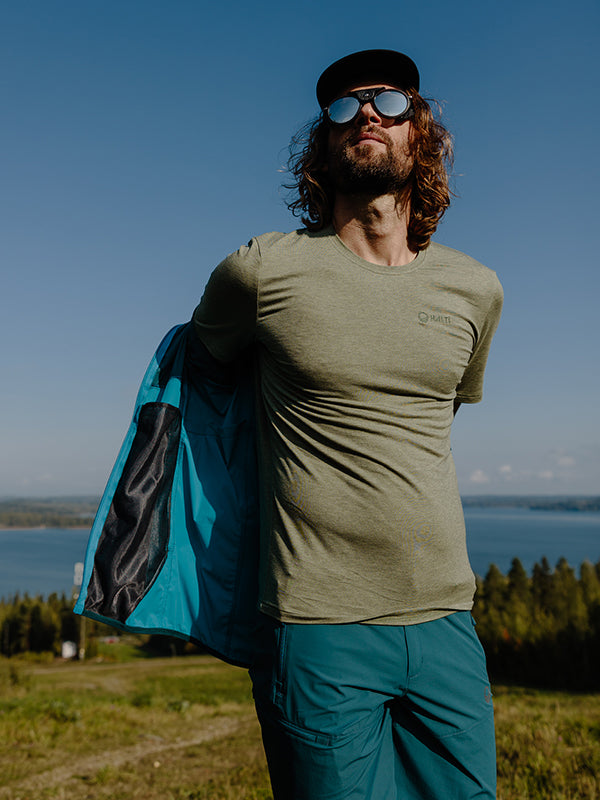 Aurinkolasit ja lippalakki päässä oleva mies pukee sinisen Halti Tuntu II Merino T-paita Miesten -paidan ulkona järven ja puiden rannalla.