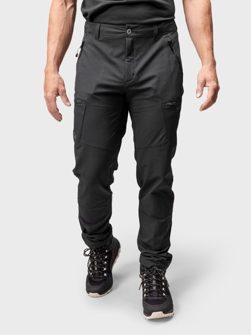 Halti Pallas Evo Brushed X-stretch miesten ulkoiluhousuihin ja mustiin vaelluskenkiin pukeutunut henkilö seisoo vaaleanharmaan tausta-asun vieressä.