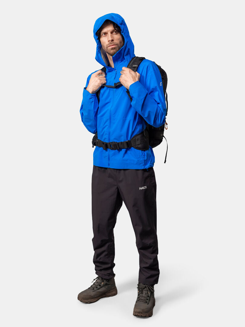 Mies, jolla on sininen Halti Pisarat 2,5L DX Kuoritakki, mustat housut, saappaat ja reppu, seisoo kasvot menosuuntaan.