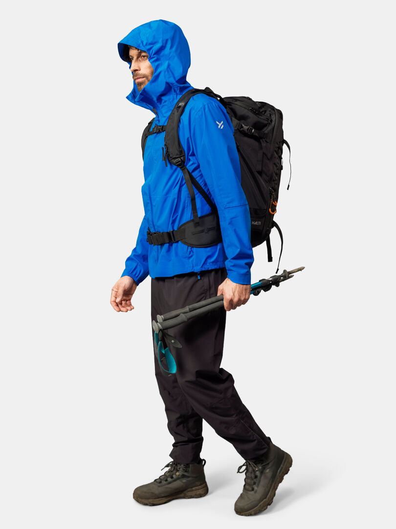 Siniseen Halti Pisarat 2,5L DX Kuoritakki Miesten, mustiin housuihin ja saappaisiin pukeutunut henkilö seisoo reppu ja vaellussauvat mukanaan.