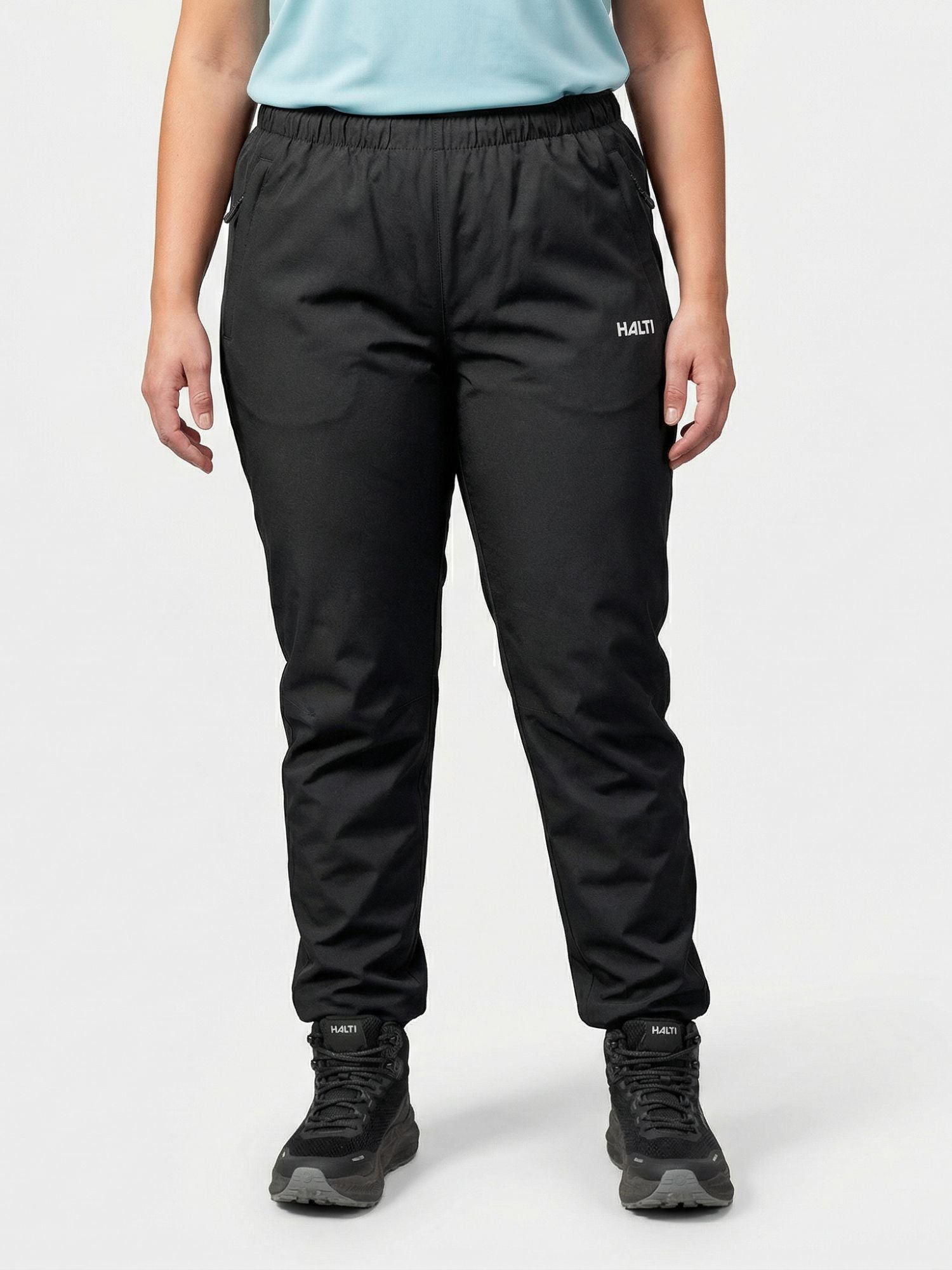 Henkilö seisoo Halti Forter DX Kuorihousut Naisten Plus, mustat lenkkarit ja vaaleansininen paita valkoisella pohjalla.