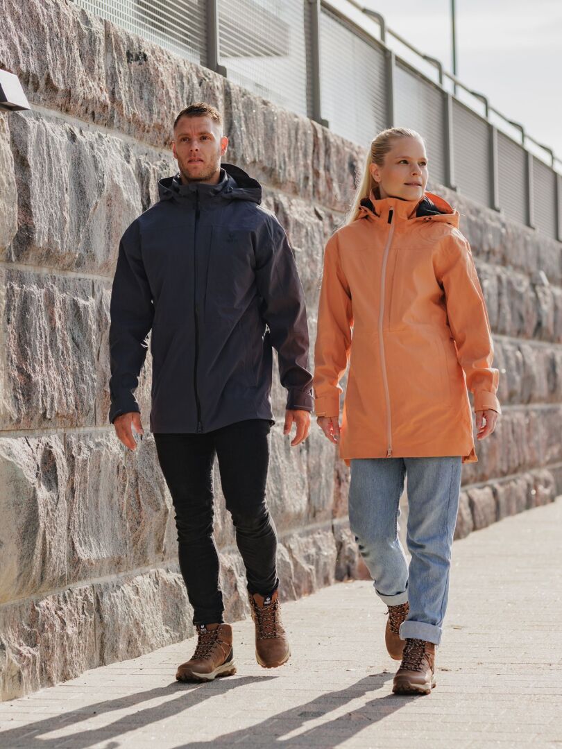 Mies mustassa takissa ja nainen Halti Kallas DX Parkatakki Naisten takissa kävelevät kivimuurin ohi aurinkoisena päivänä.