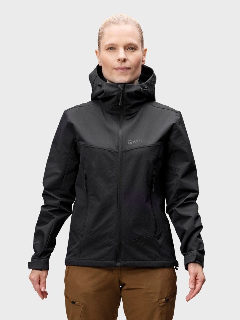 Henkilö, jolla on Halti Pallas Evo Brushed X-stretch Takki Naisten ja ruskeat housut, seisoo vaaleanharmaata taustaa vasten.