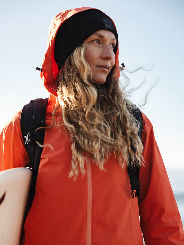Henkilö, jolla on pitkät aaltoilevat hiukset, oranssi Halti Staala DX Kuoritakki Naisten ja musta pipo pitää surffilautaa rannalla.