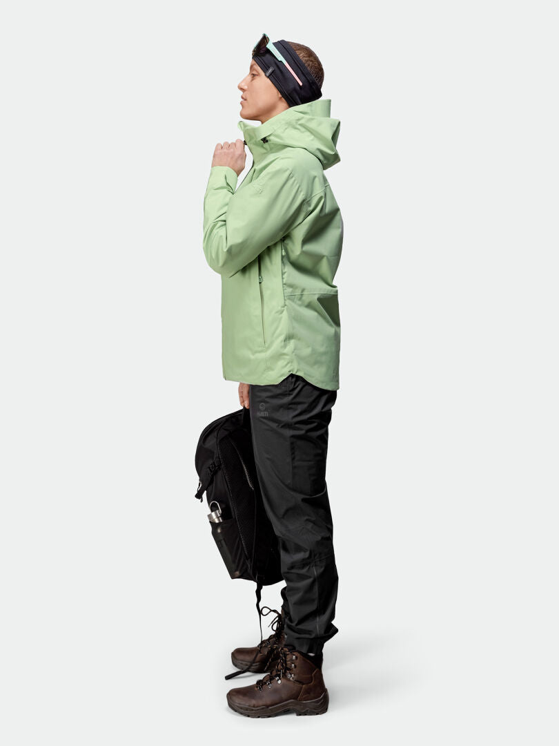 Henkilö, jolla on Halti Staala DX Kuoritakki Naisten, mustat housut ja saappaat, seisoo profiilissa pitelemässä mustaa reppua.