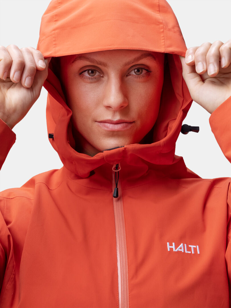 Oranssiin Halti Staala DX Kuoritakki Naisten huppu ylös vedettynä, kameraan päin tavallista taustaa vasten.