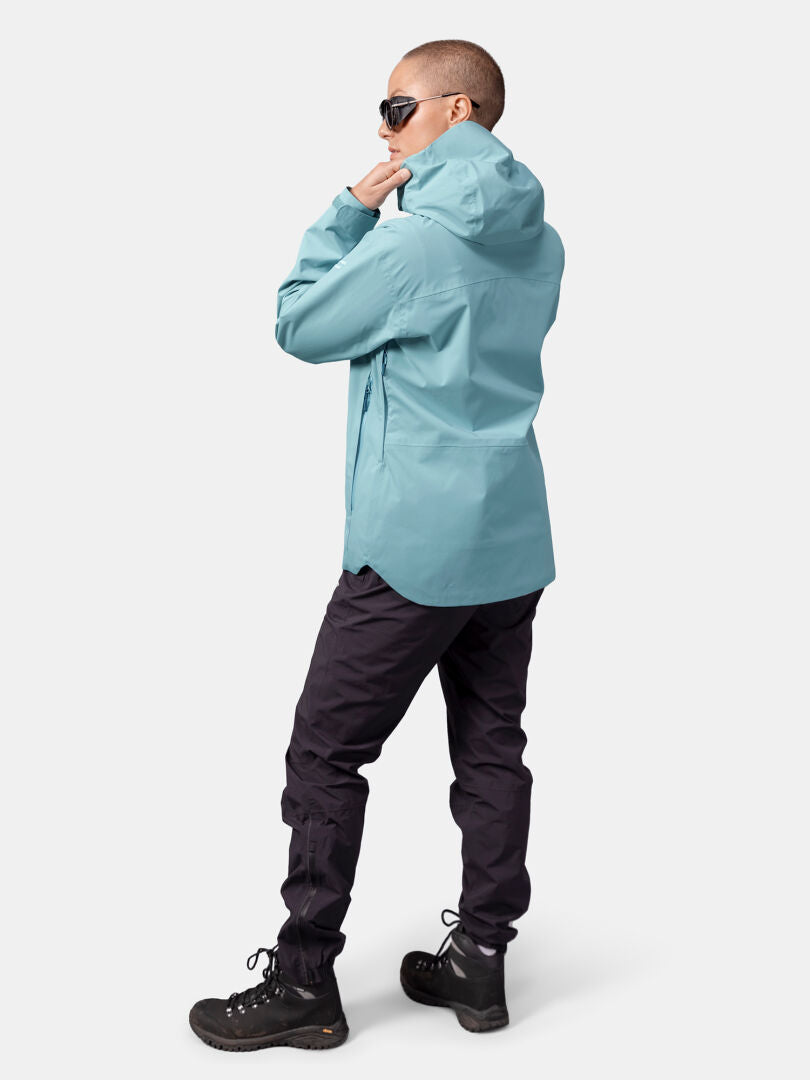 Henkilö, jolla on yllään Halti Staala DX Kuoritakki Naisten, mustat housut ja mustat vaelluskengät, seisoo ja katsoo poispäin.