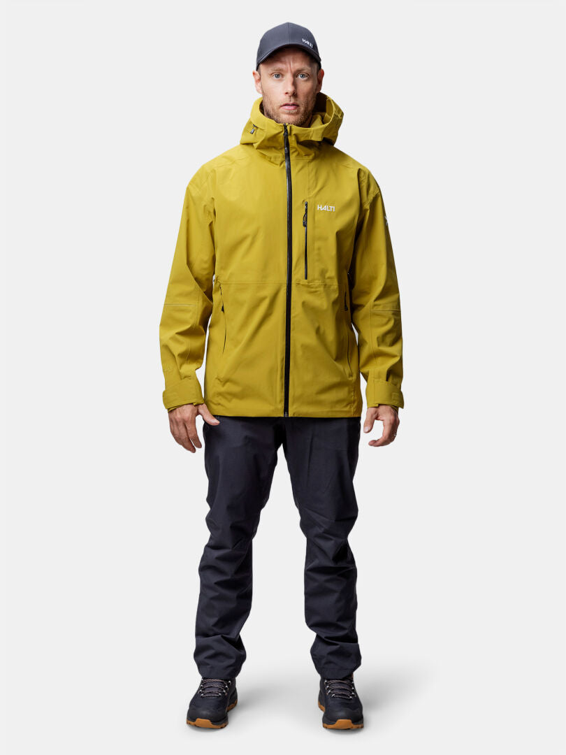 Valkoista taustaa vasten seisoo mies, jolla on keltainen Halti Staala DX Kuoritakki Miesten, tummat housut ja mustat kengät.