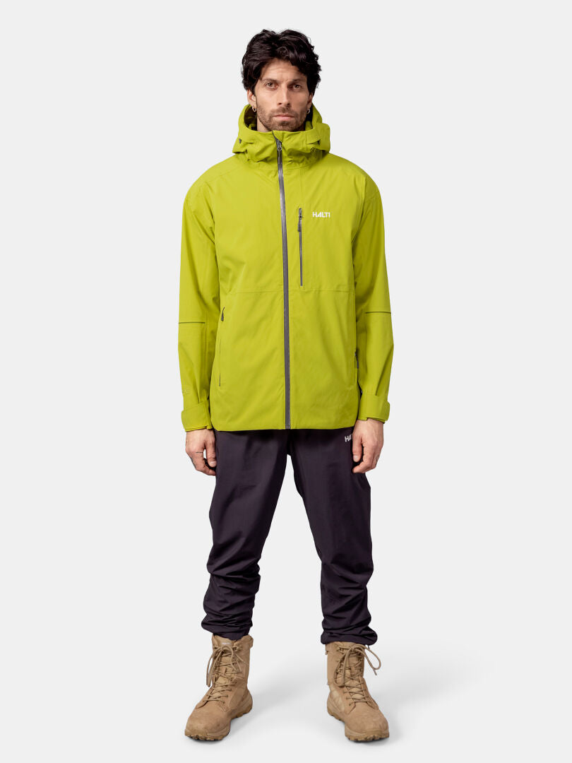 Mies katsoo eteenpäin limenvihreässä Halti Staala DX Kuoritakki Miesten, mustissa housuissa ja ruskehtavissa saappaissa tavallista taustaa vasten.