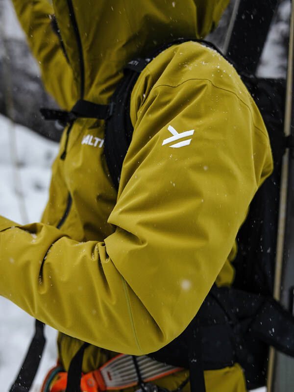 Halti Staala DX Kuoritakki Miesten henkilö seisoo ulkona lumessa, reppu selässä ja varusteita kantamassa.