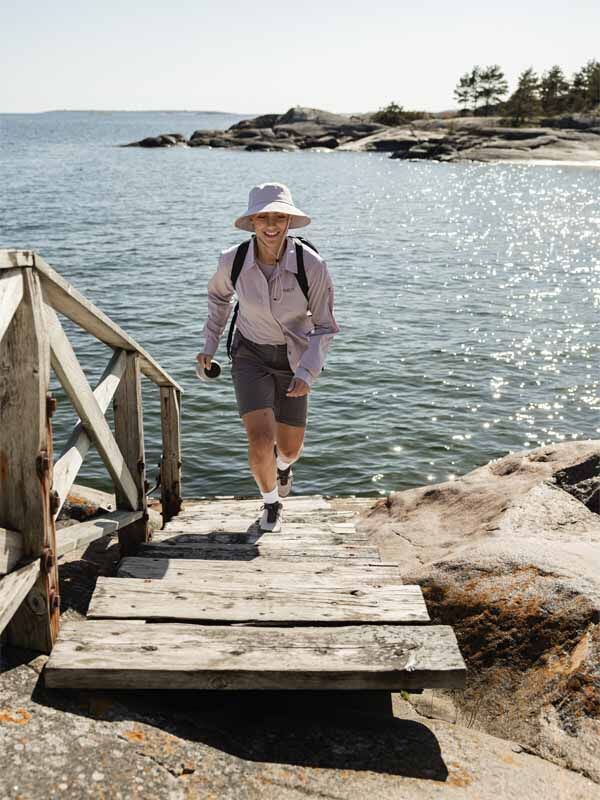 Henkilö, jolla on yllään Haltin Pallas Cool Stretch Zip-off Housut Naisten, vaeltaa puisia portaita ylös kalliorannalta aurinkoisena päivänä.