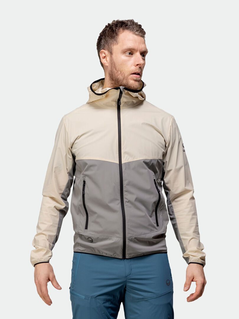 Mies seisoo vasemmalla yksinkertaista taustaa vasten yllään beigen ja harmaan Halti Pallas Cool X-stretch Takki Miesten -takki ja siniset housut.