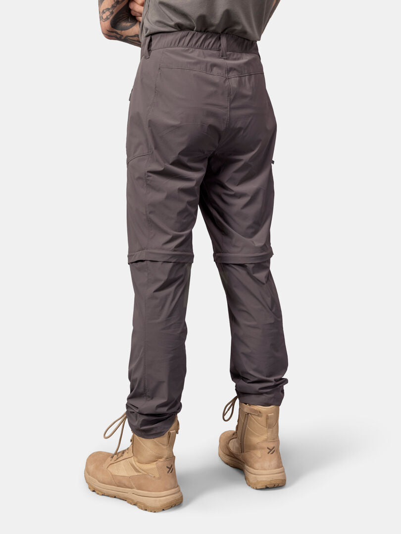 Henkilöllä on Halti Pallas Cool Stretch Zip-off Housut Miesten ja ruskeat nauhakengät valkoisella pohjalla.