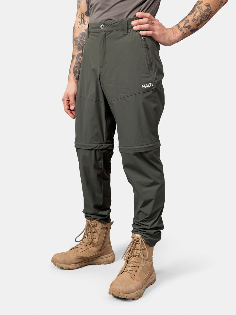 Henkilöllä on yllään Halti Pallas Cool Stretch Zip-off Housut Miesten, ruskehtavat saappaat ja harmaa paita, toinen käsi lantiolla.
