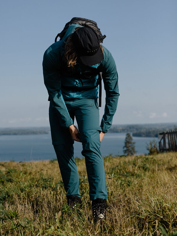 Halti Pallas Cool X-stretch Zip-off -housuihin pukeutunut mies oikaisee housujensa lahkeita järven rannalla puiden ja ruohon keskellä.