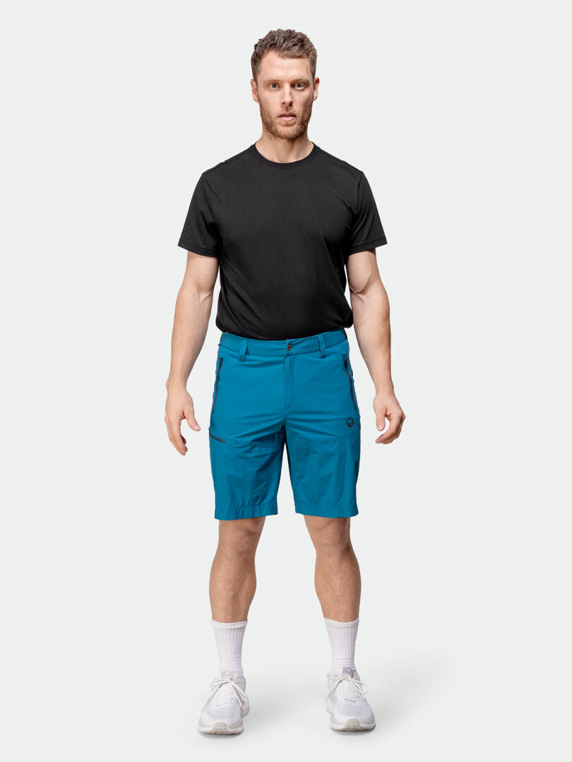 Mies kasvot eteenpäin Halti Pallas Cool X-stretch -shortseissa, mustassa t-paidassa, valkoisissa sukissa ja lenkkareissa yksiväristä taustaa vasten.