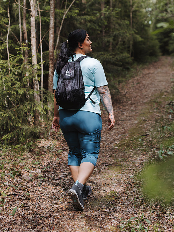 Hän kävelee metsäpolulla rehevän vehreyden keskellä Halti Pallas Cool X-stretch Caprit Naisten Plus -vaatteissa, joissa on UPF 90+.