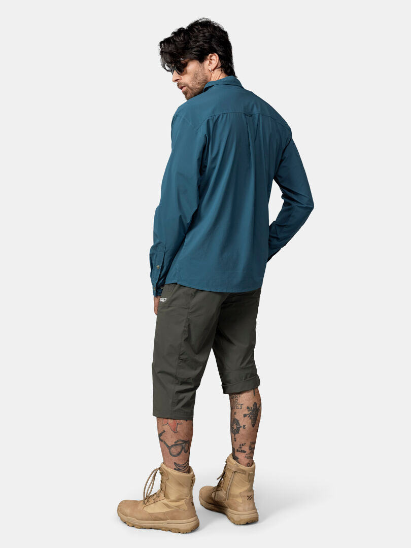 Mies katsoo poispäin sinisessä Halti Samoy pitkähihainen kauluspaita, harmaat shortsit, ruskettuneet saappaat ja jalkatatuoinnit.