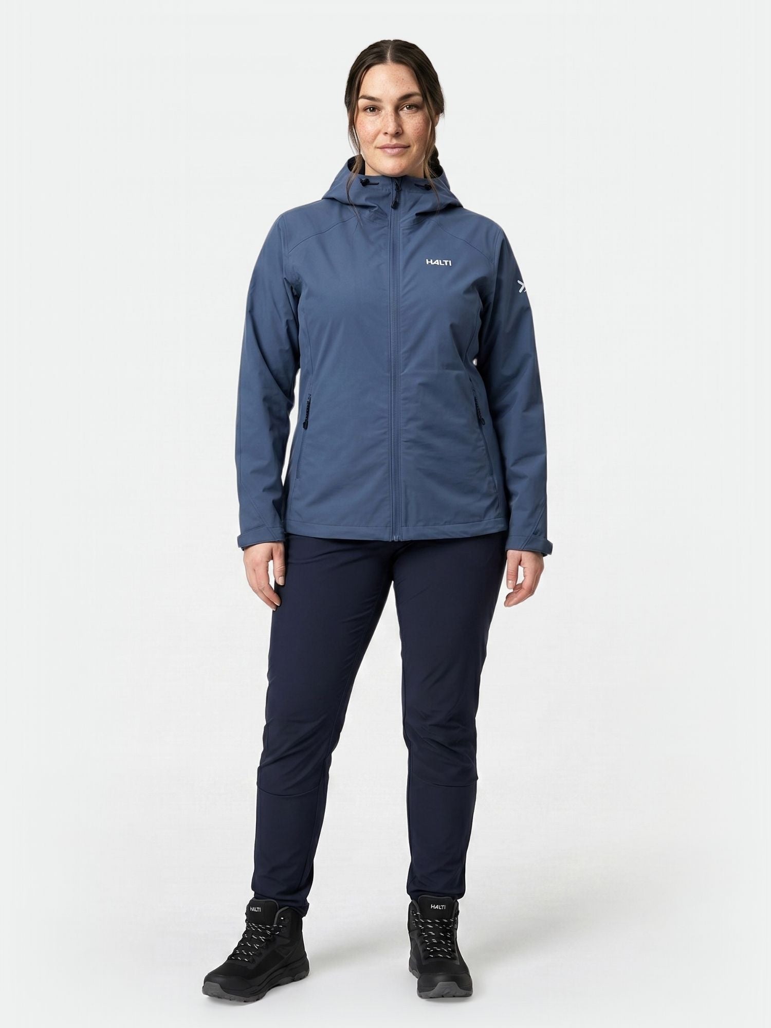 Henkilö seisoo kasvot eteenpäin päin pukeutuneena Halti Finholma X-stretch Takki Naisten Plus -takkiin, tummiin housuihin ja mustiin vaelluskengiin.