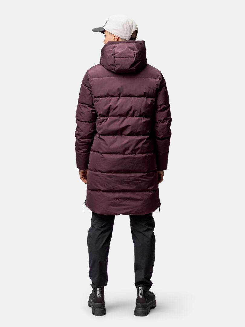 Henkilö kasvot menosuuntaan ruskeankeltaisessa Halti Linjat Parkatakki Naisten, mustat housut ja kengät, vaalea lippis, yksivärinen tausta.