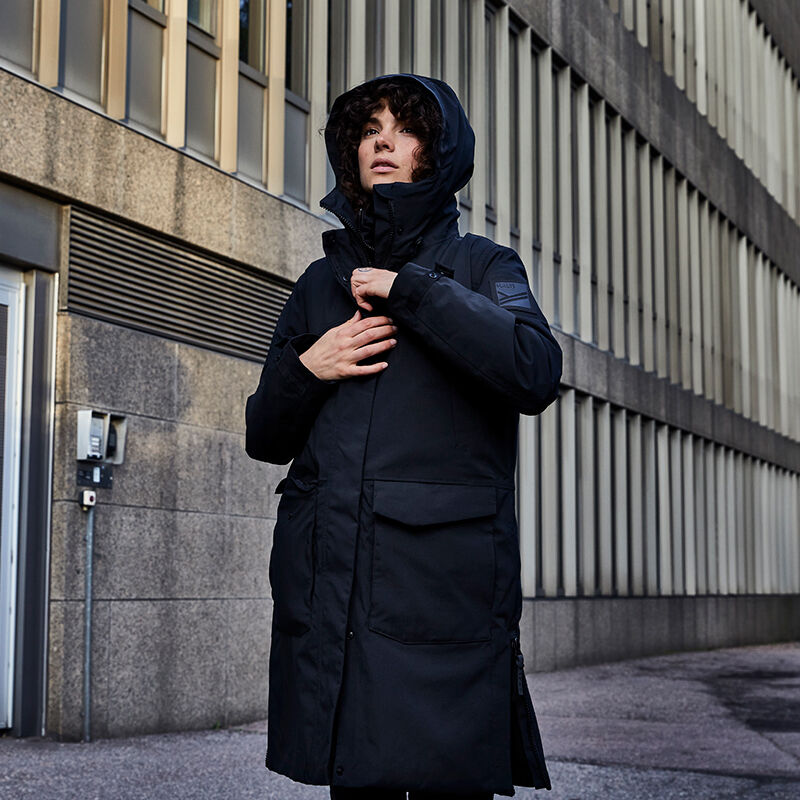 Henkilö, jolla on musta Halti Bergga DX Talvitakki Naisten seisoo ulkona modernin rakennuksen vieressä ja säätää etuvetoketjua.