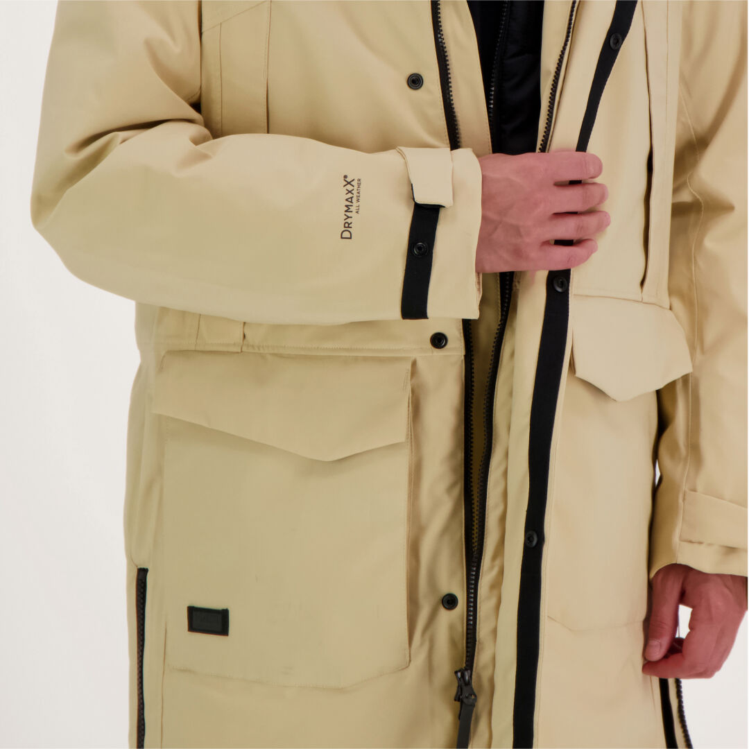 Mies vetää vetoketjun kiinni beigestä Halti Bergga DrymaxX Talvitakista, jossa on suuret taskut ja DRY-MAXX-teksti vasemmassa hihassa.