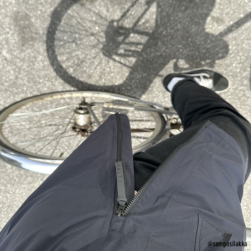 Halti Kurvi 3L DX Miesten Parkatakki ja mustat housut -hahmo seisoo polkupyörän vieressä ja heittää varjon jalkakäytävälle.