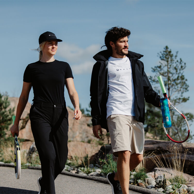 Kaksi ihmistä Halti Drive Naisten X-stretch Chinot -kengissä kävelee ulkona tennismailat kädessään ja nauttii aktiivisesta liikunnasta.