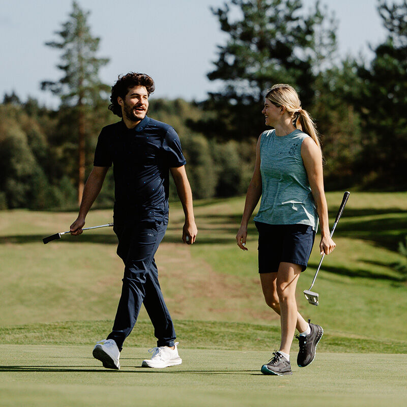 Mies ja nainen kävelevät puiden ja ruohon ympäröimällä golfkentällä Halti Drive Miesten X-stretch Chinotissa.