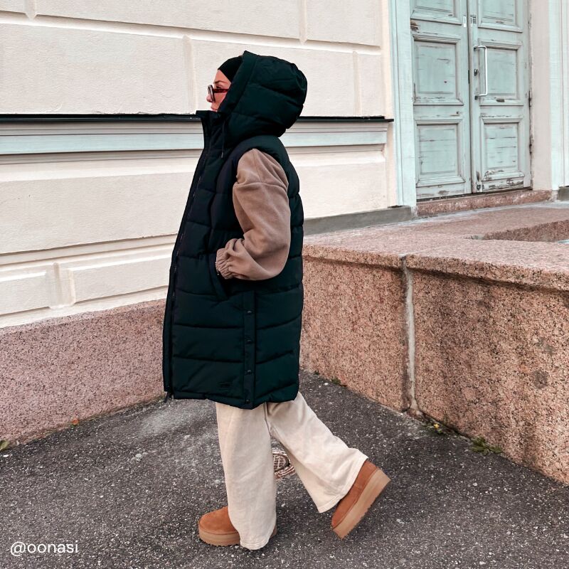 Henkilö, jolla on musta Halti Penger Toppaliivi Naisten, beiget housut ja ruskeat saappaat, kävelee vaalean rakennuksen ohi.