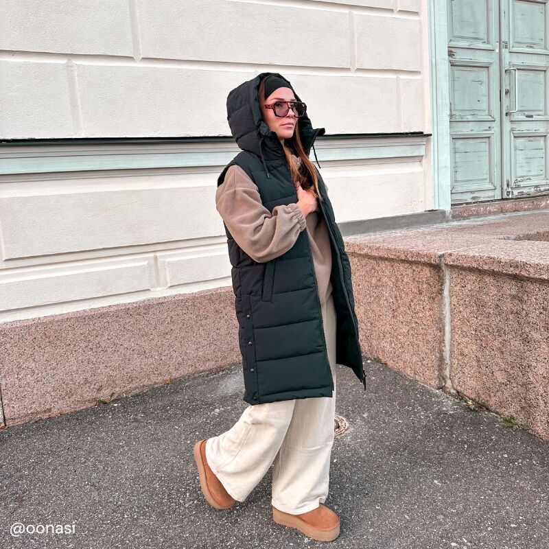 Mustaan Halti Penger Toppaliivi Naisten ja beigeihin housuihin pukeutunut henkilö kävelee vaalean rakennuksen ohi.