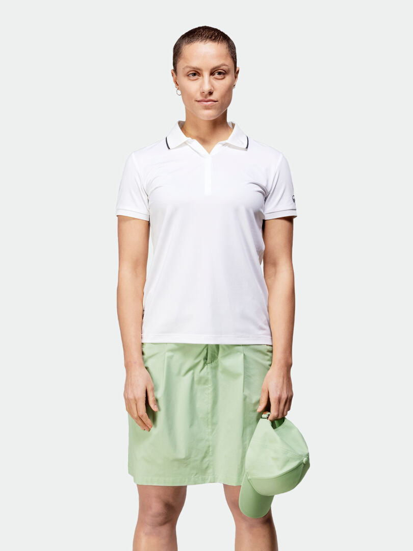 Nainen valkoisessa poolopaidassa ja Halti Drive X-stretch -shortsihameessa seisoo vihreä lippalakki päässään valmiina ulkoiluun.