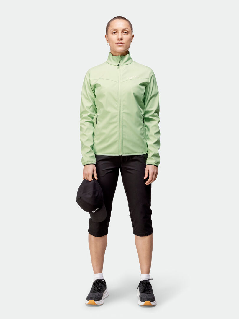 Henkilö Halti Wedge Softshell-takki Naisten farkuissa, mustissa caprit ja lenkkareissa seisoo ja pitelee mustaa lippalakkia.