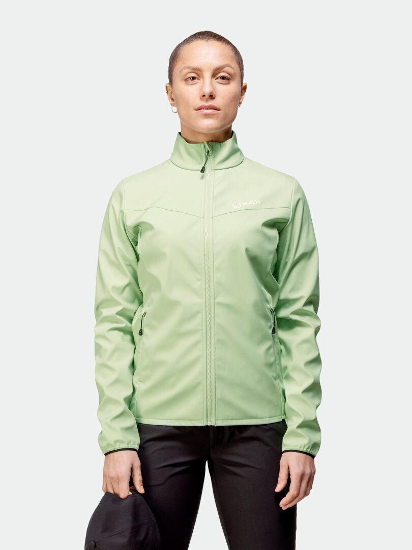 Henkilö seisoo kasvot eteenpäin pukeutuneena Halti Wedge Softshell-takki Naisten takkiin ja mustiin housuihin ja pitelee mustaa esinettä.