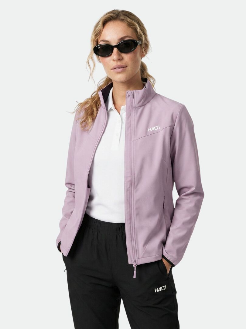 Aurinkolaseihin, Halti Wedge Softshell-takki Naisten ja mustiin housuihin pukeutunut henkilö seisoo kädet taskuissa tavallista taustaa vasten.