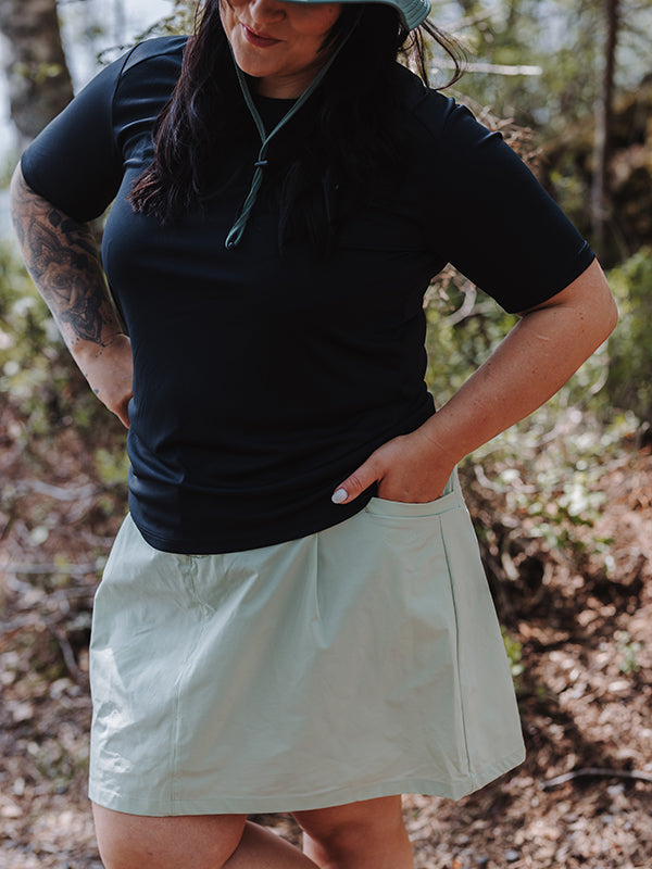 Henkilö ulkona mustassa paidassa ja Halti Drive X-stretch -shortseissa Naisten Plus -housuissa, tyylikkäässä plus-koon hameessa, kädet taskuissa.