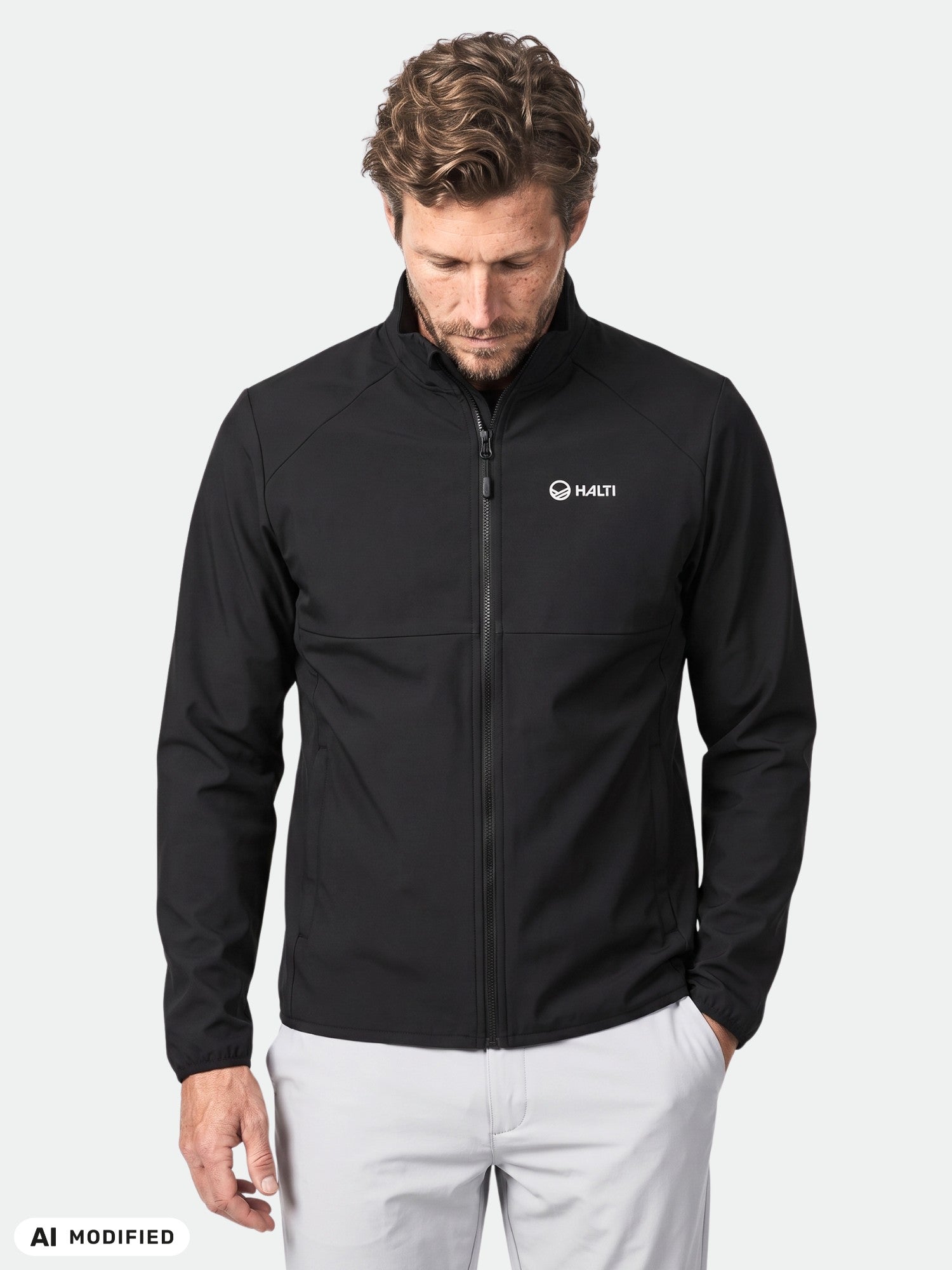 Mies, jolla on yllään Halti Wedge Softshell -takki (kierrätysmateriaalia) ja valkoiset housut, harmaalla taustalla.