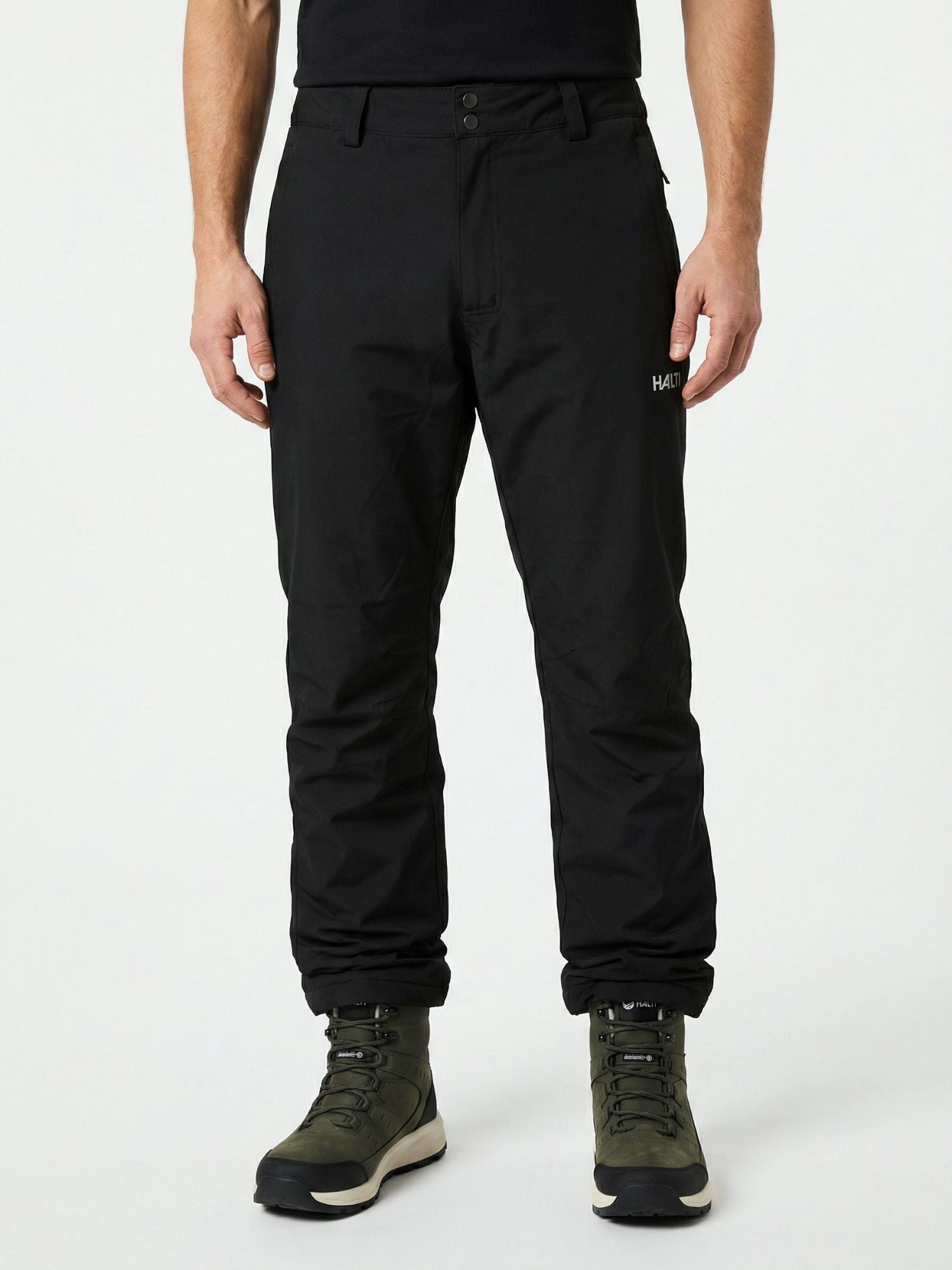 Halti Routa Toppahousut Miesten ja vihreisiin vaelluskenkiin pukeutunut henkilö seisoo tavallista vaaleaa taustaa vasten.