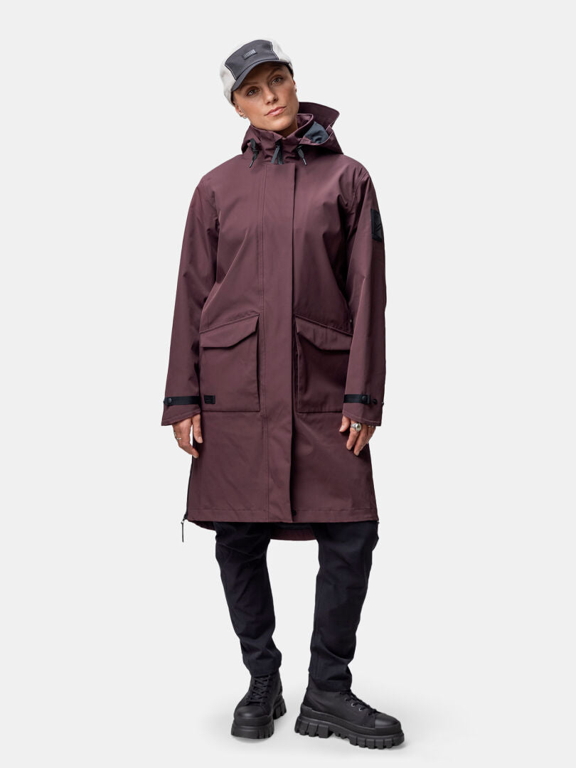 Henkilö, jolla on ruskeankeltainen Halti Tokoi II DX Parkatakki Naisten, mustat housut, paksut saappaat ja harmaa lippis, seisoo kasvot menosuuntaan.